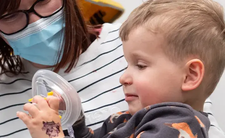 A woman wearing a mask helps a young boy hold a medical mask.